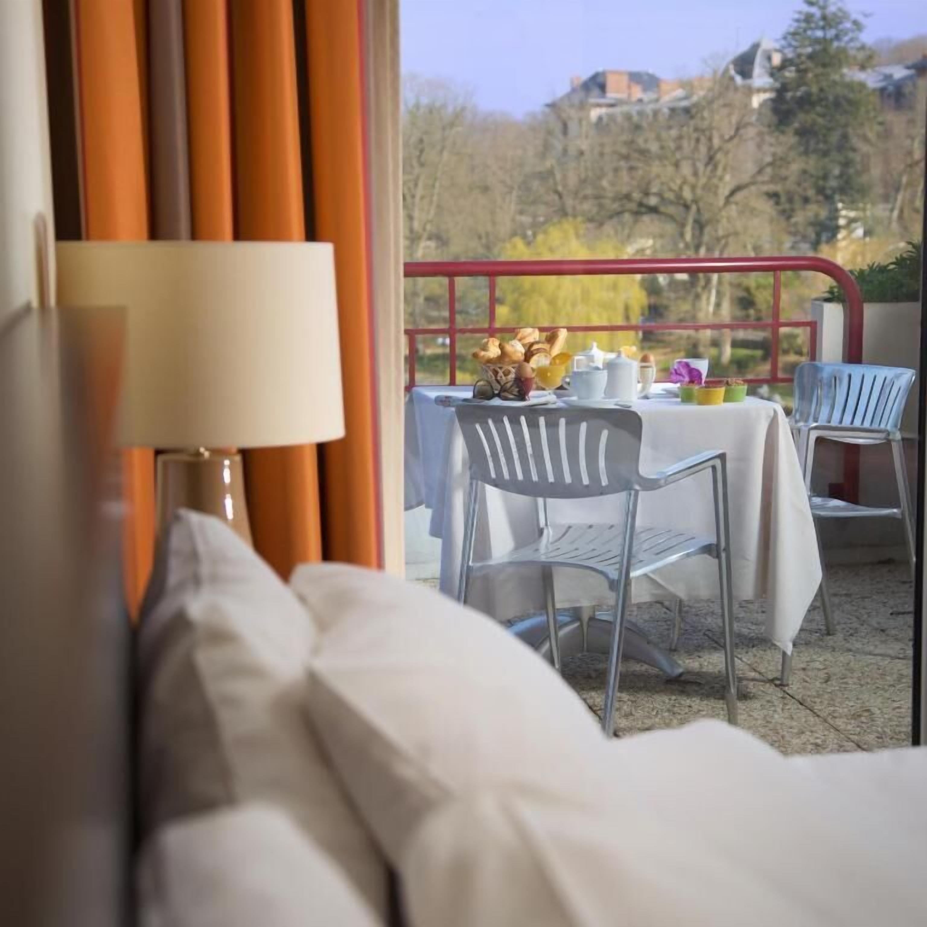 A balcony with a table set for breakfast, a chair, and a view of trees and buildings.