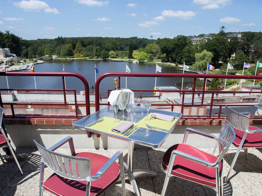 An outdoor dining area with tables and chairs, overlooking a body of water with flags and a bridge in the background.
