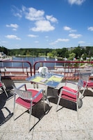 An outdoor dining area with tables and chairs, overlooking a body of water with flags and a bridge in the background.