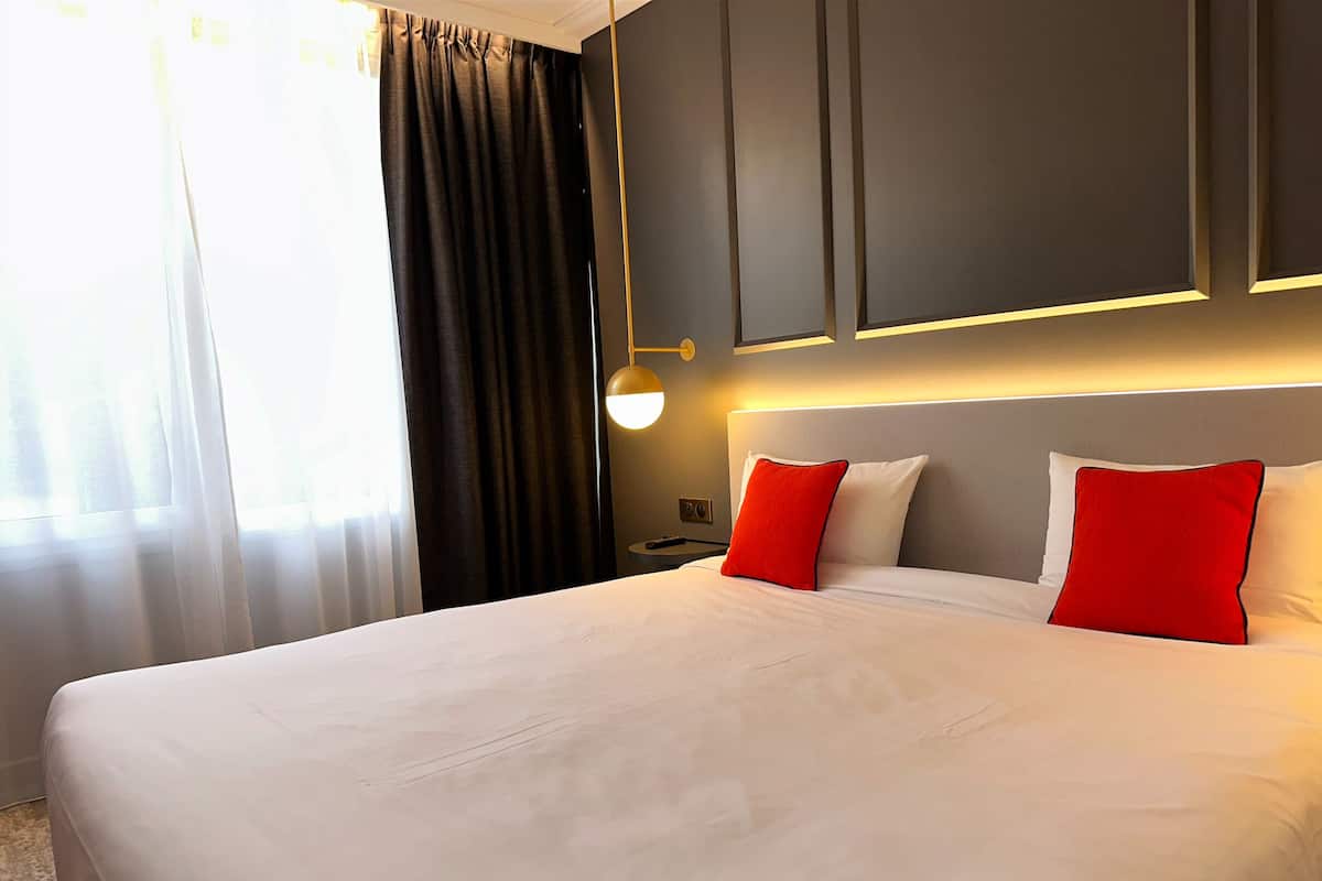Een hotelkamer met een groot bed, donkere gordijnen, een nachtkastje en een wandlamp.