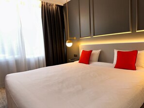Een hotelkamer met een groot bed, donkere gordijnen, een nachtkastje en een wandlamp.