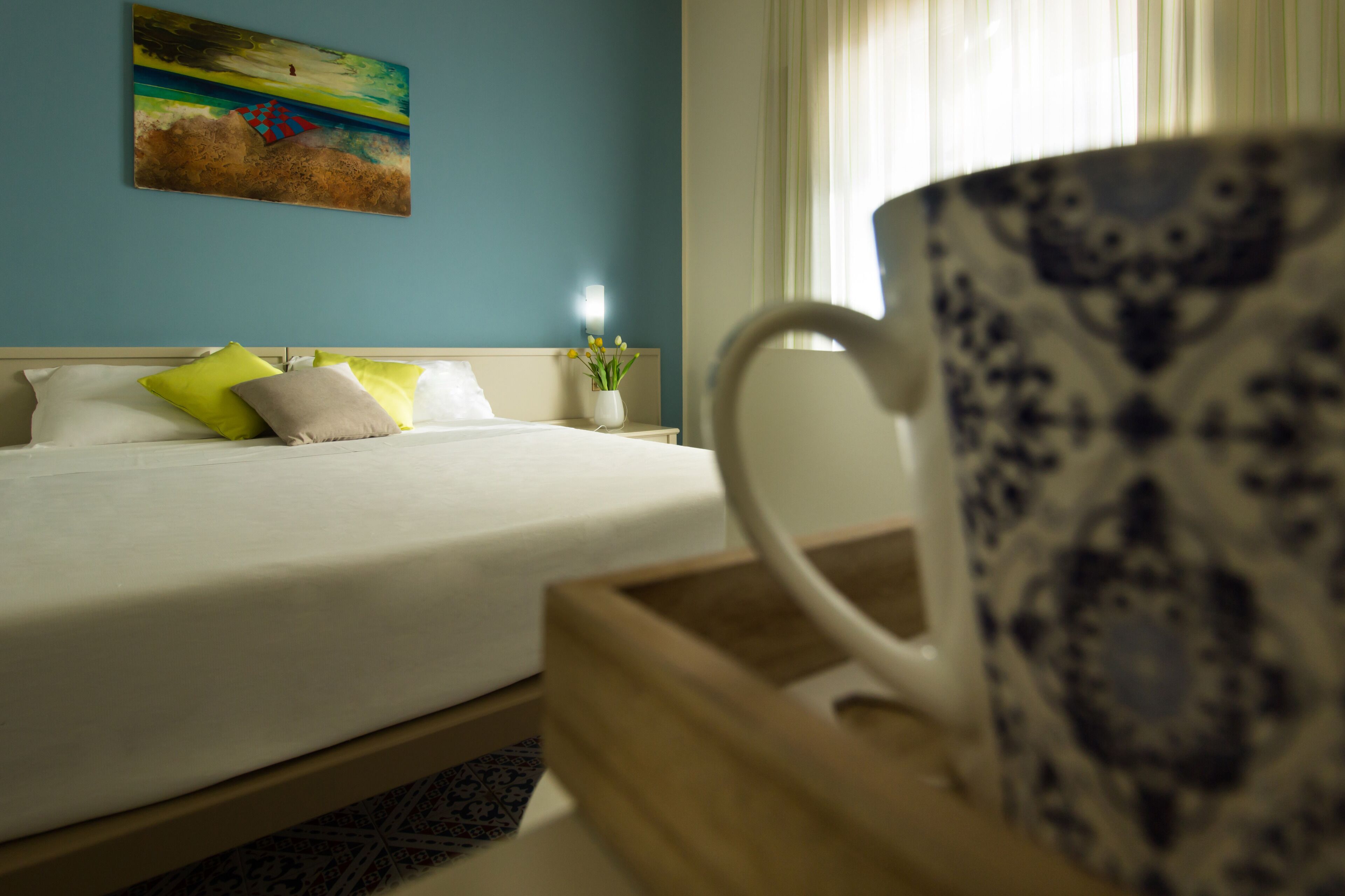 Hotellihuone, jossa on sänky, tyynyjä, maljakko kukkineen, taulu seinällä ja kuppi puisen tarjottimen päällä.