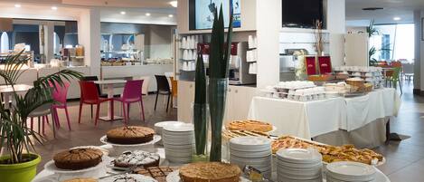 Uma área de buffet com diversos doces e pratos, rodeada por cadeiras e mesas.