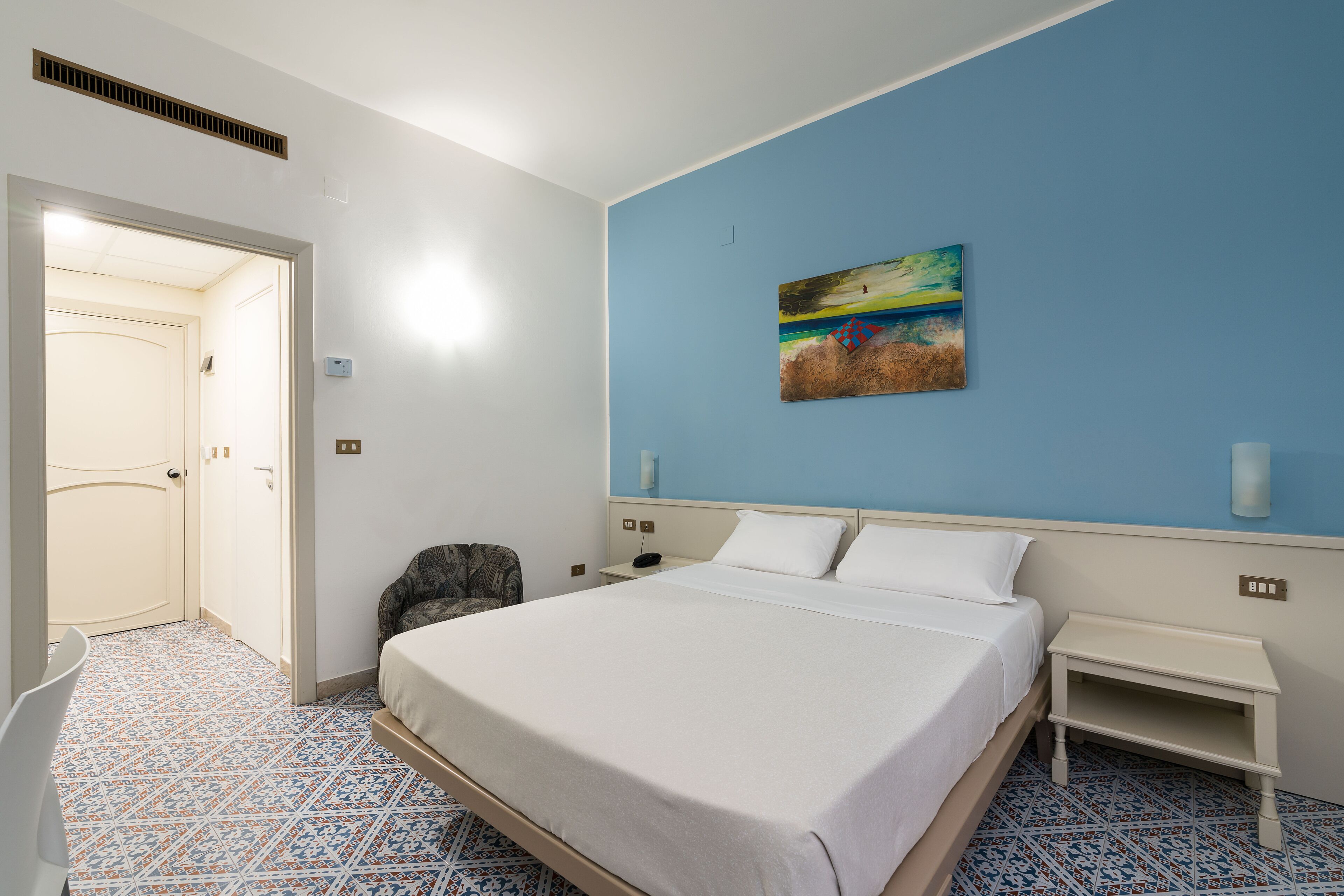 A hotel room with a bed, a chair, a nightstand, a painting on the wall, and a patterned floor.