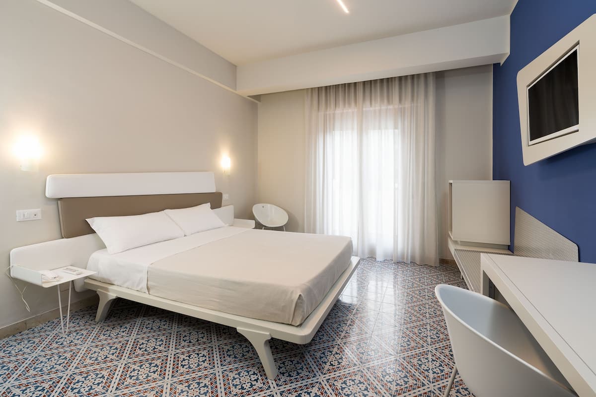 A hotel room with a bed, bedside table, chair, TV, and patterned floor.