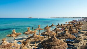 Una platja amb paraigües de palla i gandules, aigua blau turquesa i una illa a la distància.