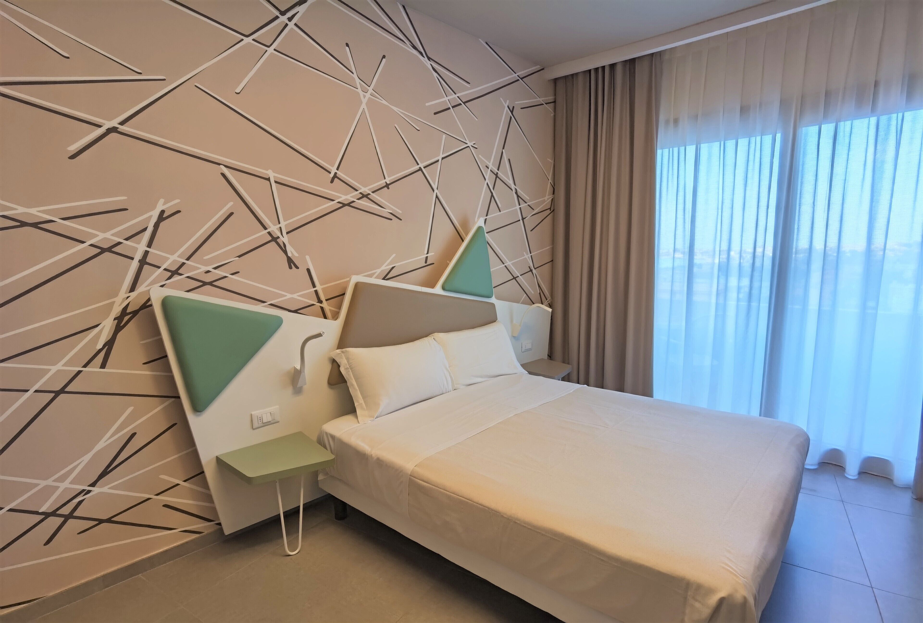 A modern hotel room with a large bed, a nightstand, a wall with abstract art, and a view of the sky through sheer curtains.