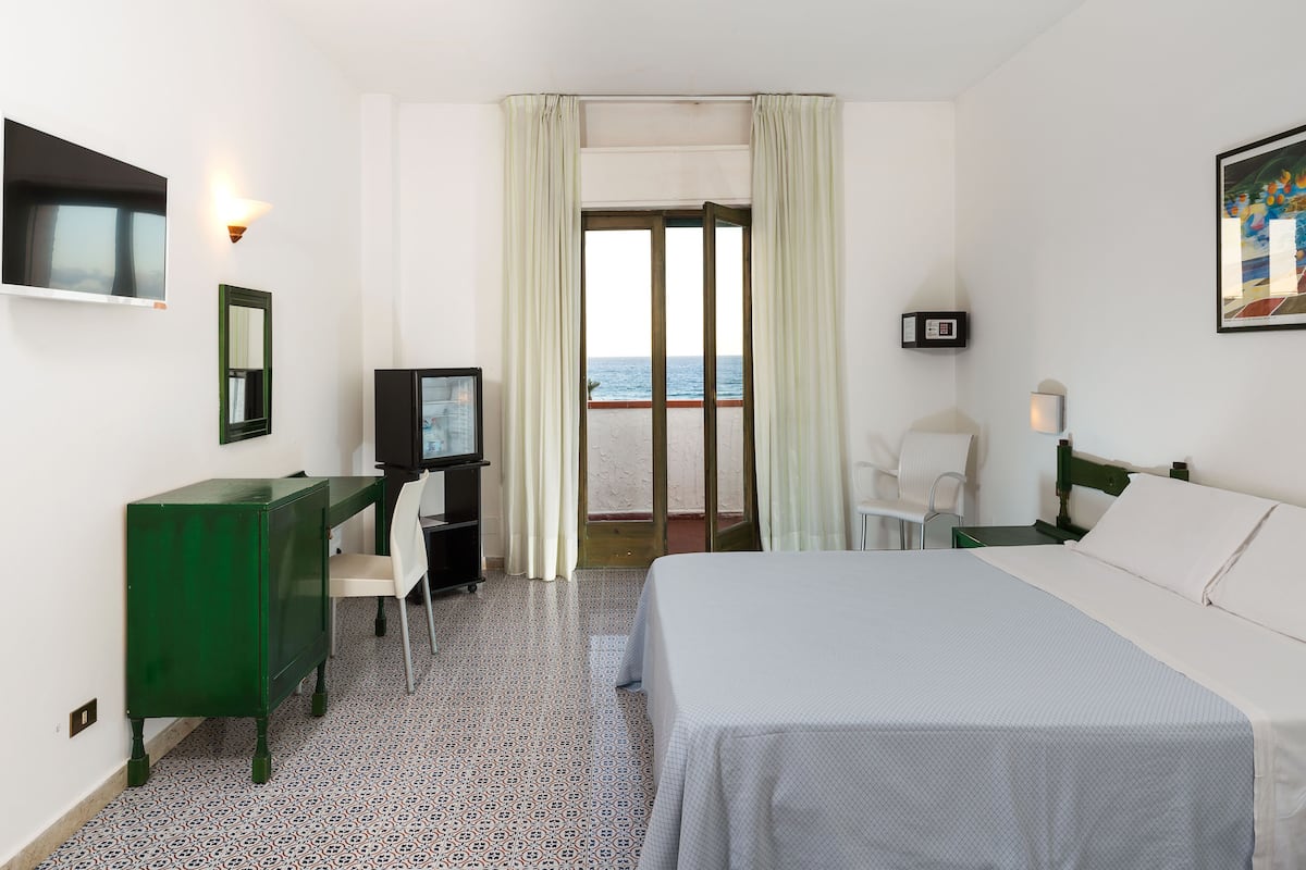 A hotel room with a bed, a desk, a chair, a TV, and a view of the sea through a large window.