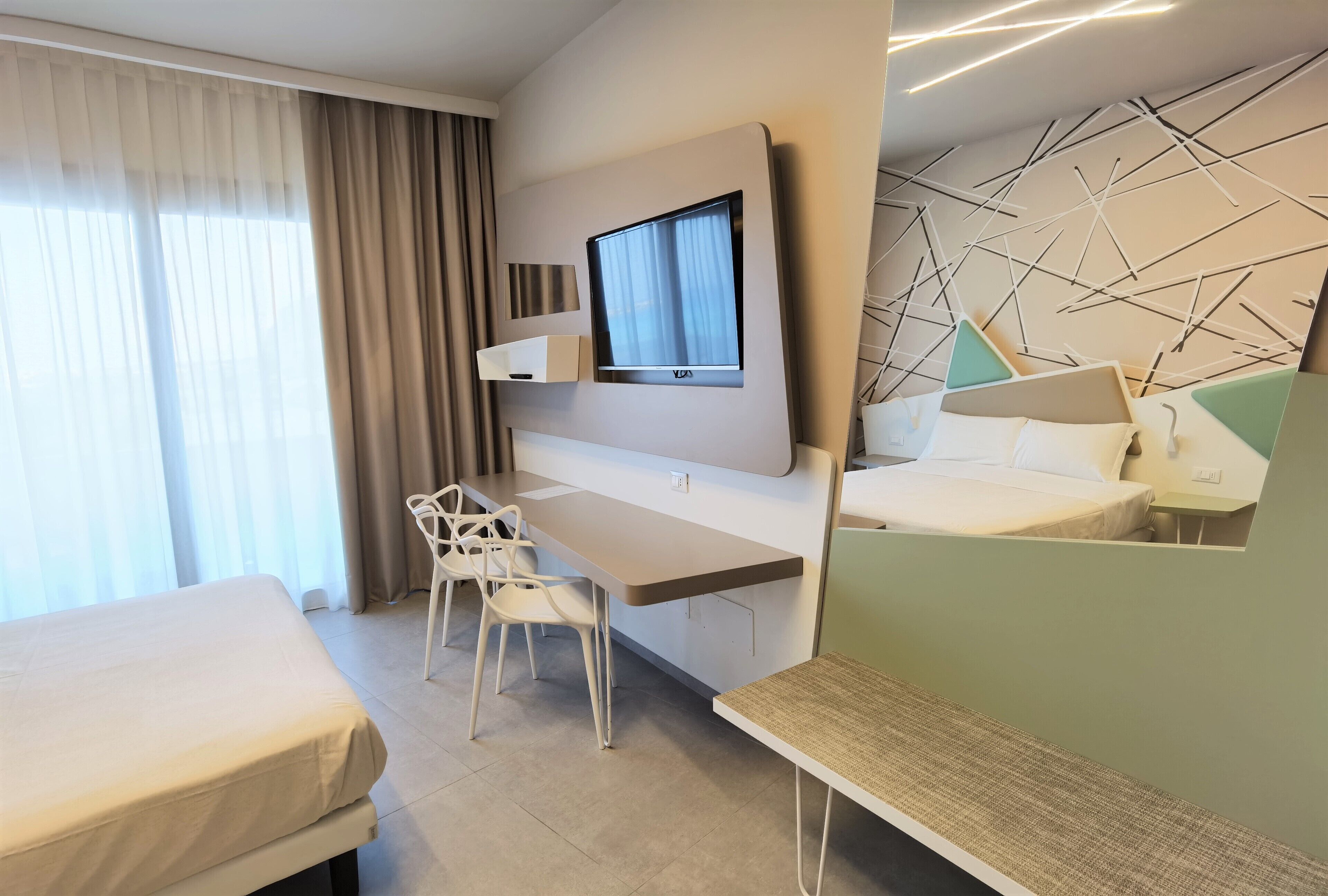 Ein modernes Hotelzimmer mit Bett, Schreibtisch, Stuhl, Fernseher und einem geometrischen Wanddesign.