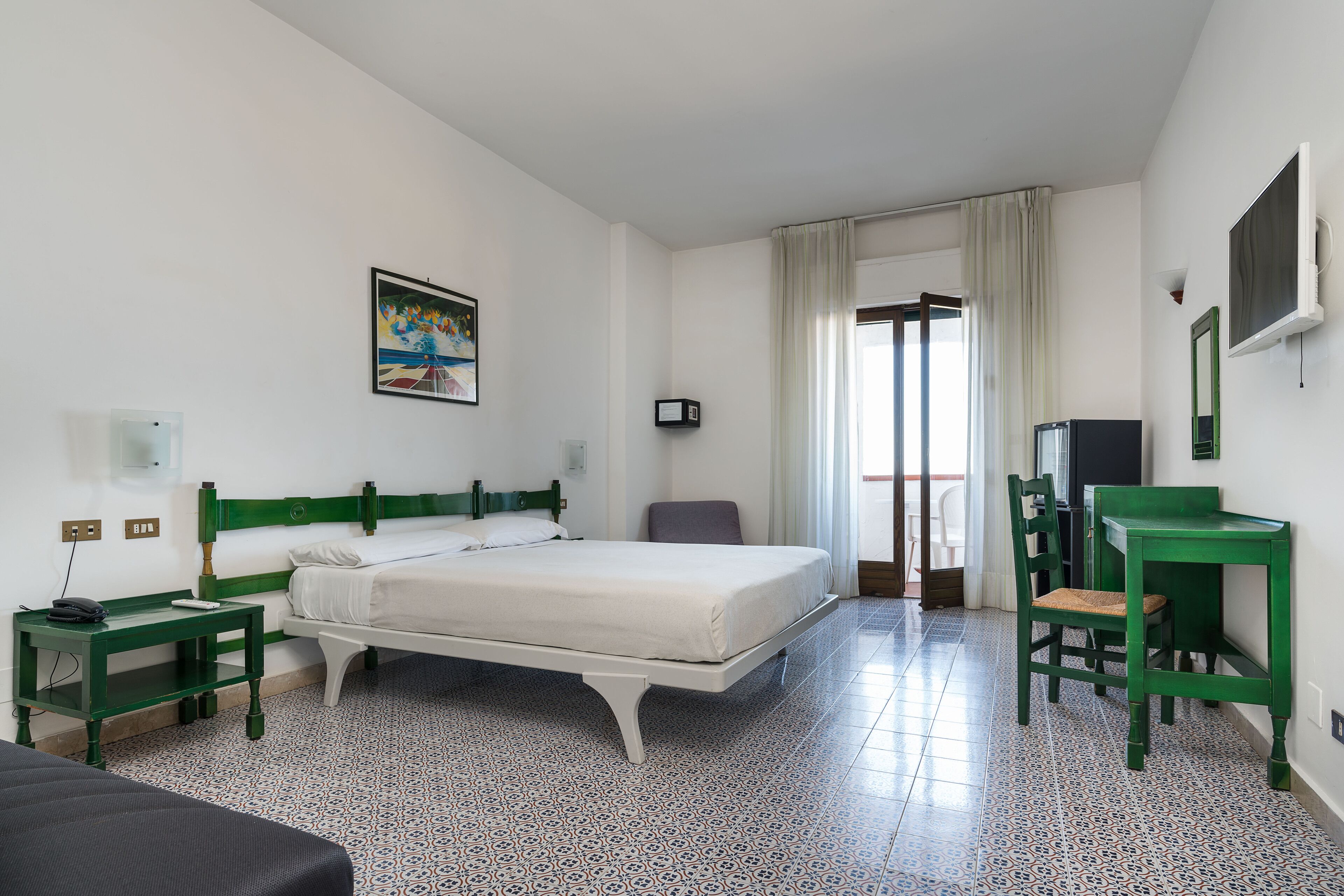 A hotel room with a bed, a desk, a chair, a TV, and a painting on the wall.