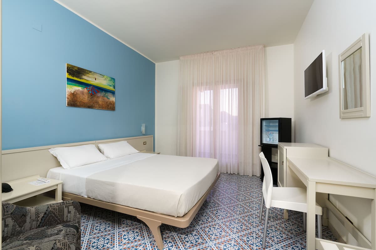 A hotel room with a bed, a desk, a chair, a TV, and a painting on the wall.