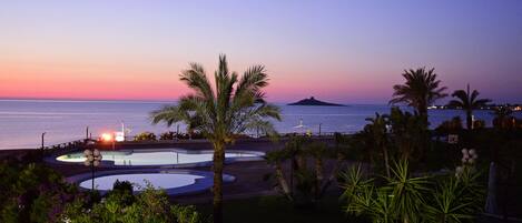 Zona de piscina con palmeras y vista al atardecer.