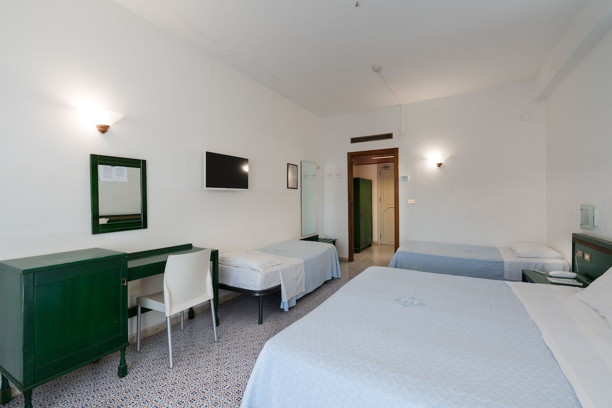 A hotel room with a large bed, a desk with a chair, two single beds, a TV, and a mirror.