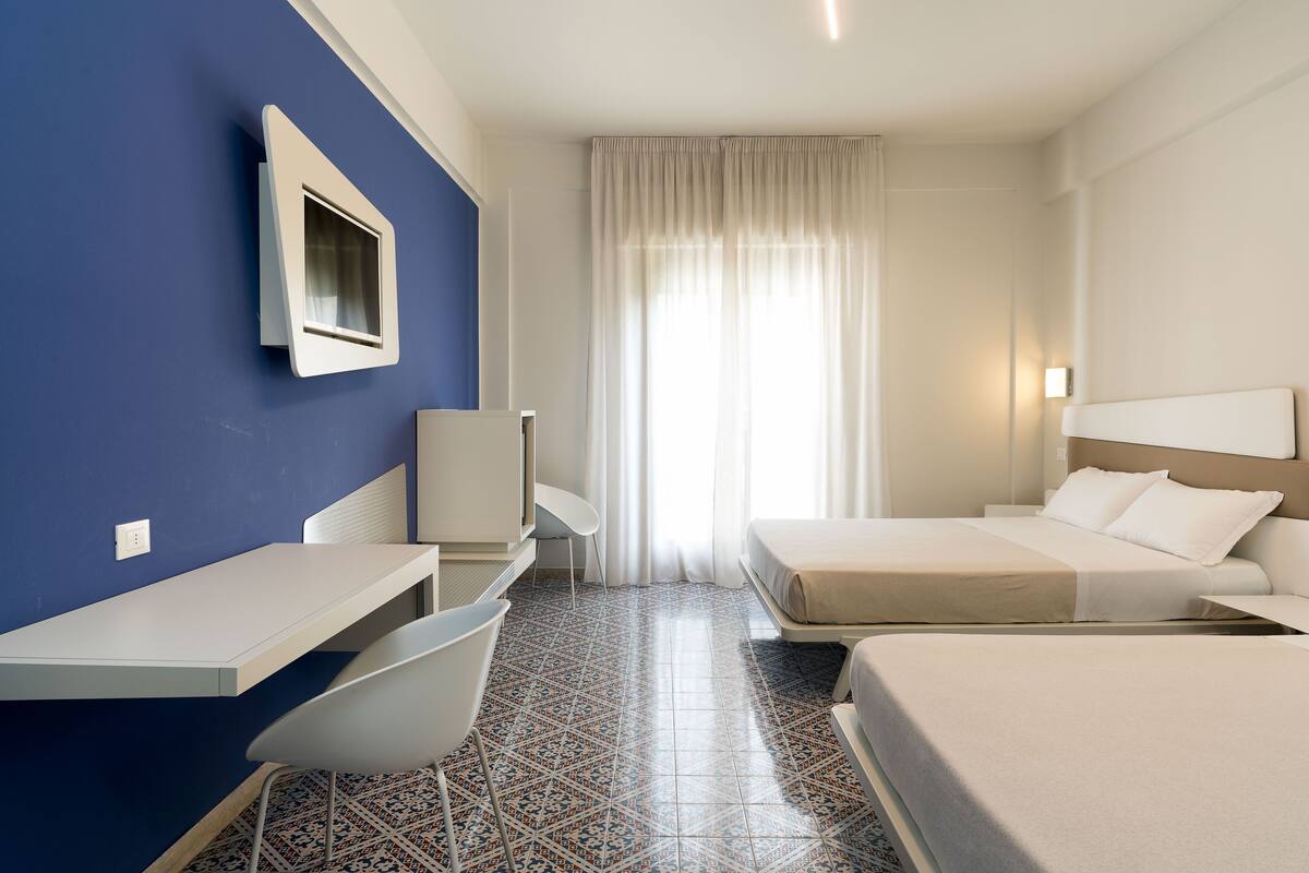 A hotel room with a desk, two beds, a chair, a TV, and a window with curtains.