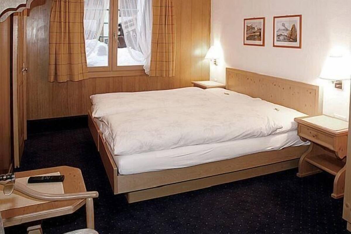 Ein Hotelzimmer mit einem hölzernen Bett, einem Schreibtisch mit Stuhl, einem Fenster mit Vorhängen und zwei gerahmten Bildern an der Wand.