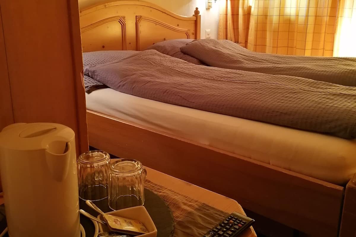 Ein Hotelzimmer mit einem hölzernen Bett, einem Nachttisch mit Fernbedienung, einer Karaffe, Gläsern und einer Schale. An der Wand hängen zwei gerahmte Bilder.
