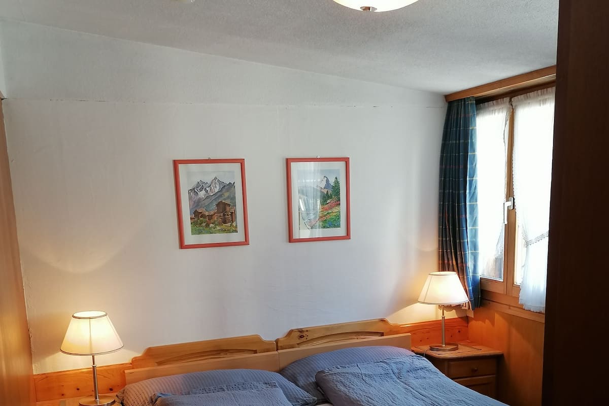 Ein ordentlich bezogenes Doppelbett mit blauer Decke, zwei Nachttische mit Lampen und zwei gerahmte Bilder an der Wand.