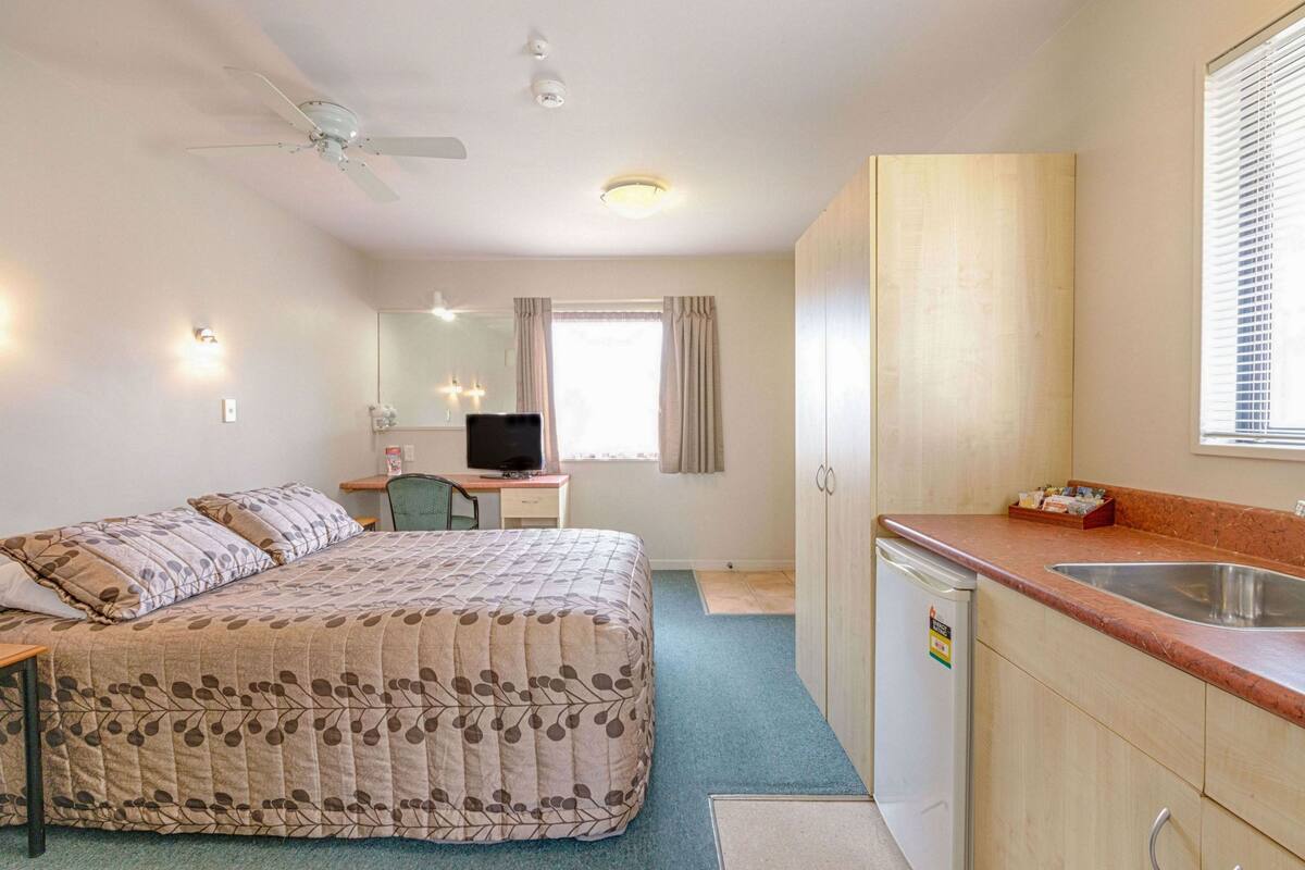A compact hotel room with a bed, a desk with a computer, a kitchenette, and a ceiling fan.