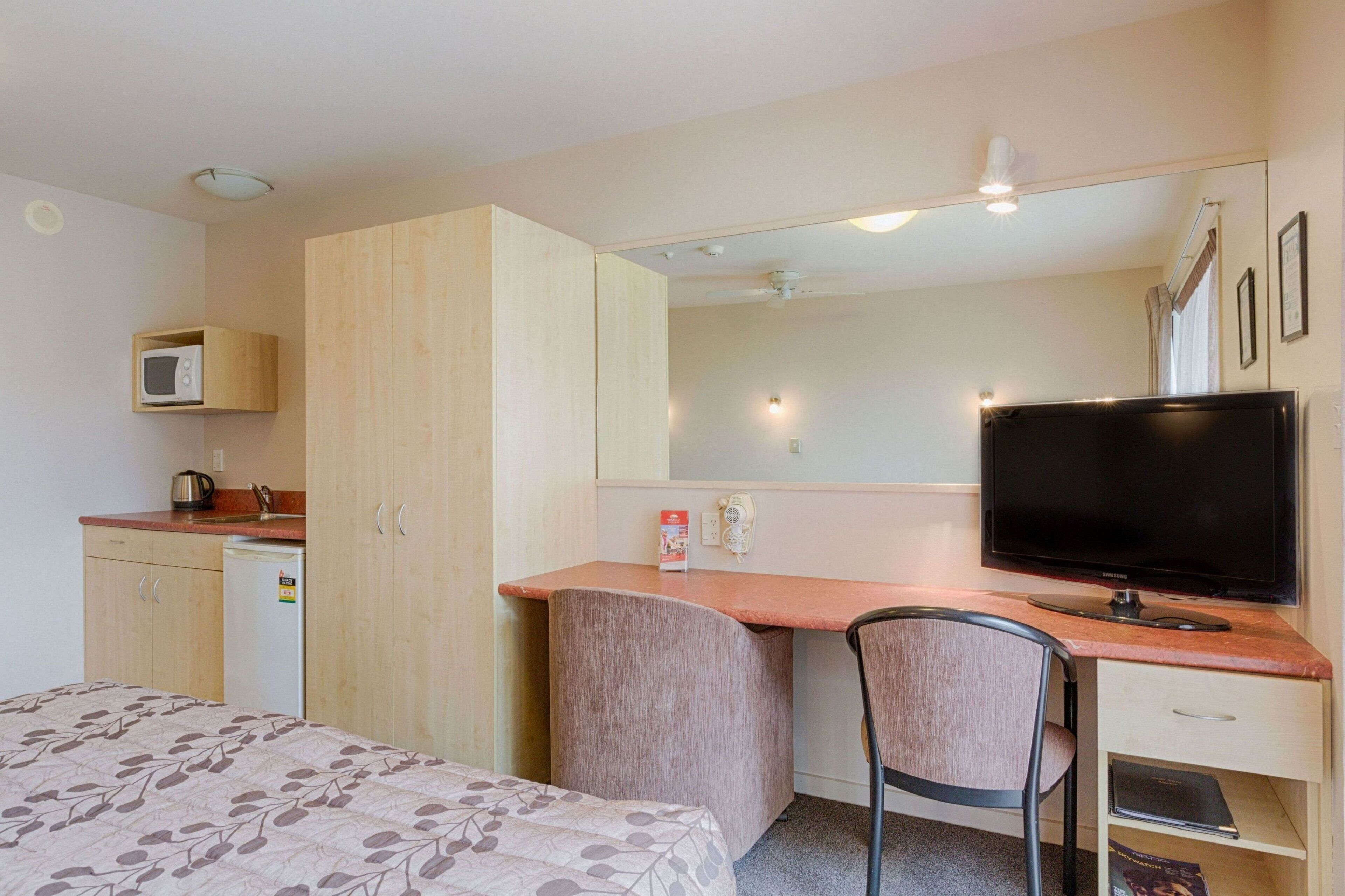 A compact hotel room with a bed, a kitchenette, a desk with a chair, a television, and a mirror.