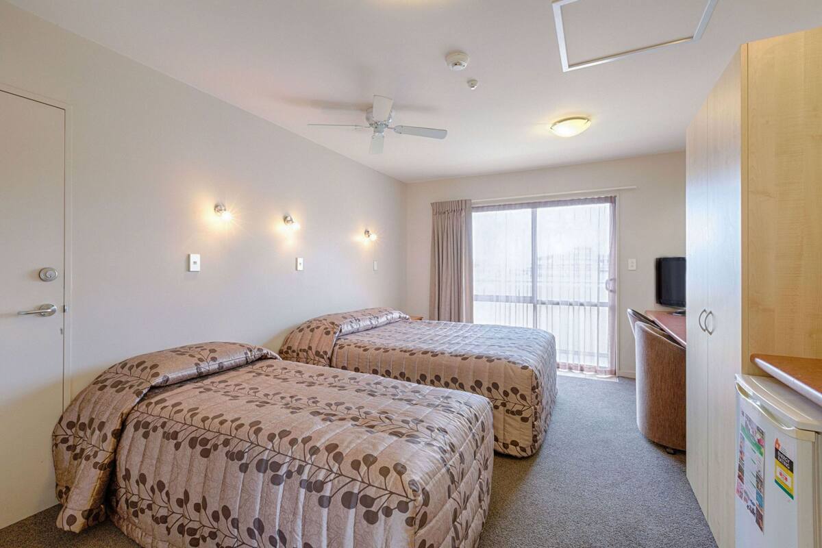 A hotel room with two beds, a ceiling fan, wall-mounted lights, a television, and a window with curtains.