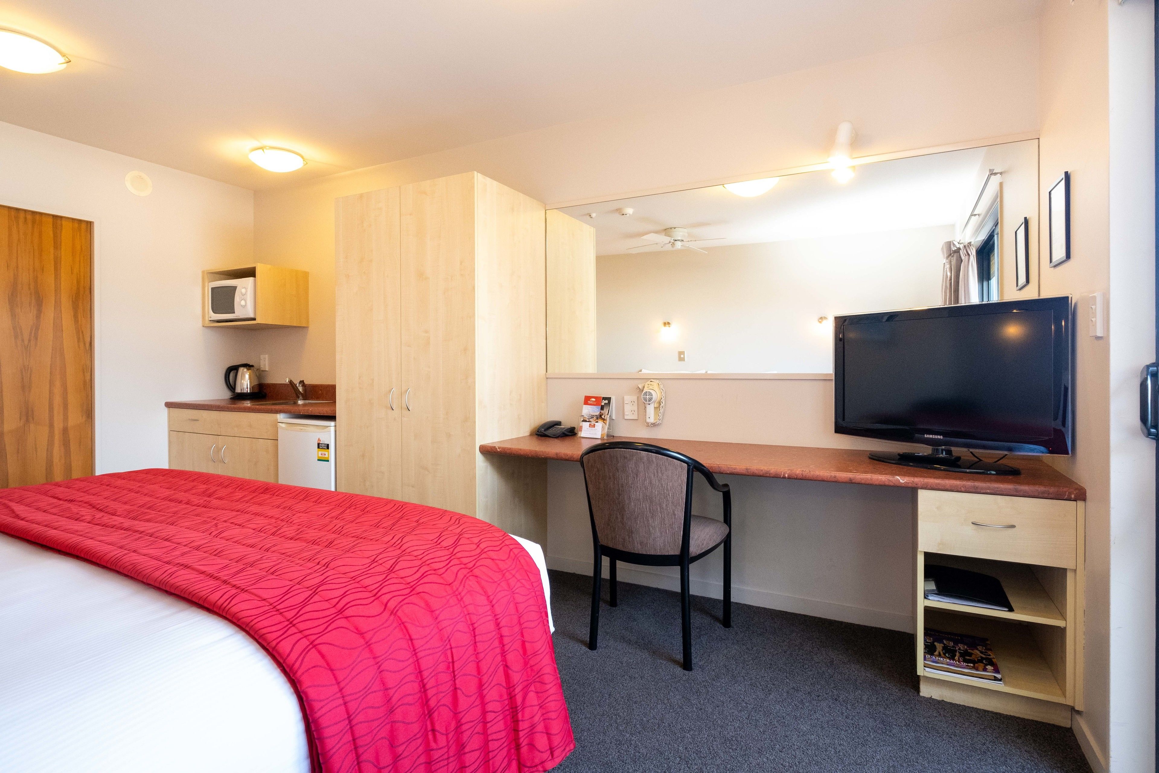 A compact hotel room with a bed, a desk, a chair, a TV, a microwave, and a small kitchenette.