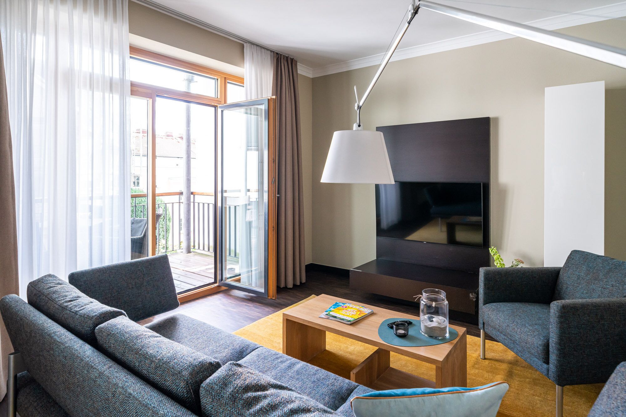 Ein modernes Wohnzimmer mit einer Couch, Sesseln, einem Couchtisch, einem Fernseher und einem Balkon mit Vorhängen.
