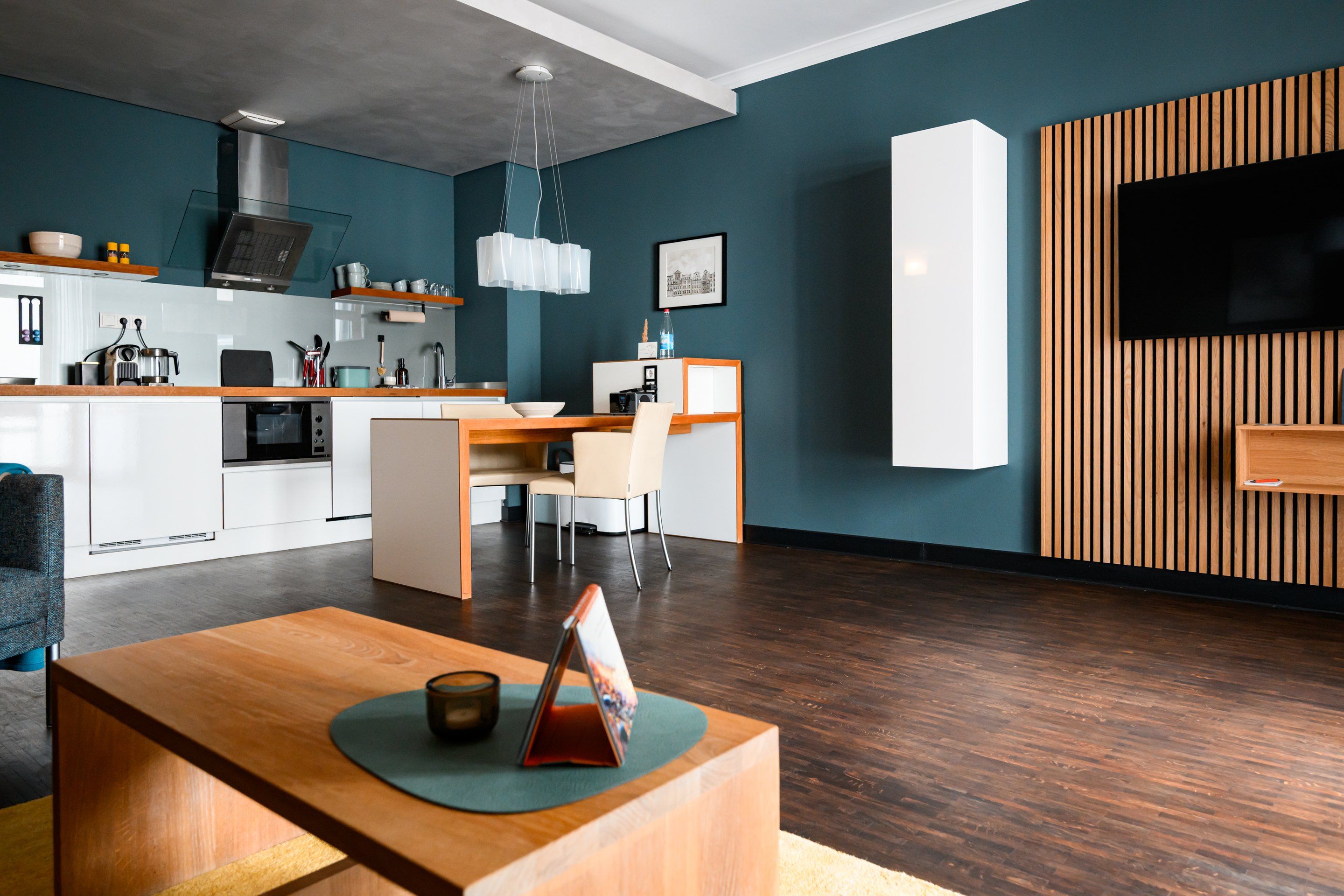 Un salon moderne avec une table basse en bois, un coin repas avec une table et des chaises blanches, une cuisine équipée d’un four à micro-ondes et d’une cuisinière, et un grand téléviseur fixé sur un mur recouvert de panneaux en bois.