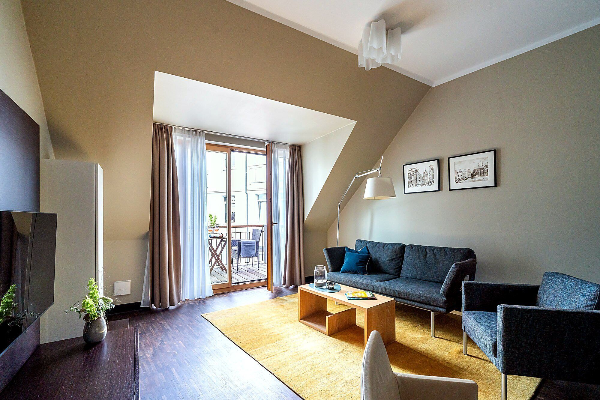 Una sala de estar moderna con un sofá, una mesa de centro y un comedor con una mesa y sillas. Hay una ventana grande con cortinas, una lámpara en el techo e imágenes enmarcadas en la pared.
