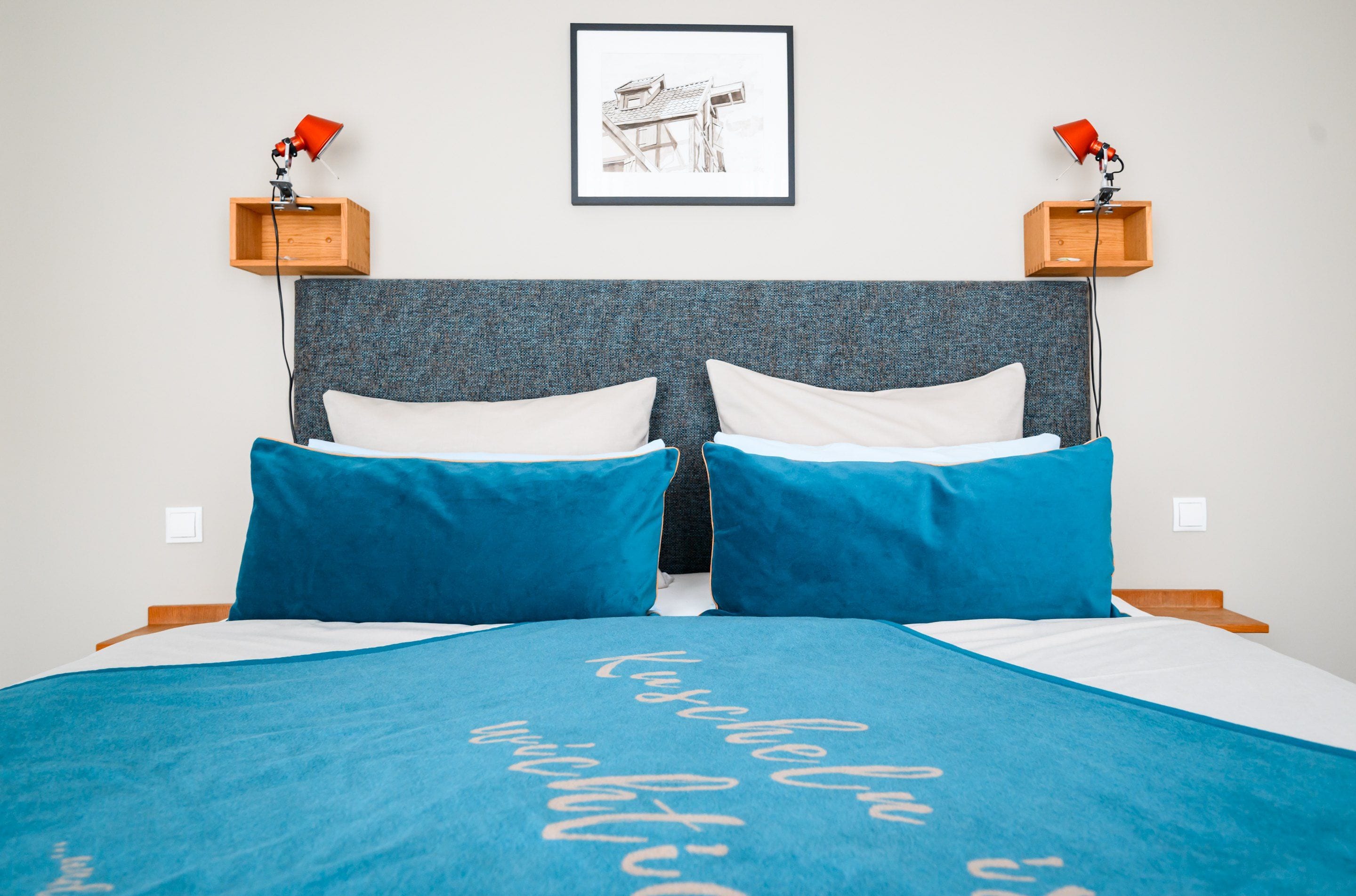 Ein Schlafzimmer mit einem großen Bett, zwei Nachttischen mit Lampen, einem gerahmten Bild an der Wand und einer blauen Bettdecke mit Aufschrift.