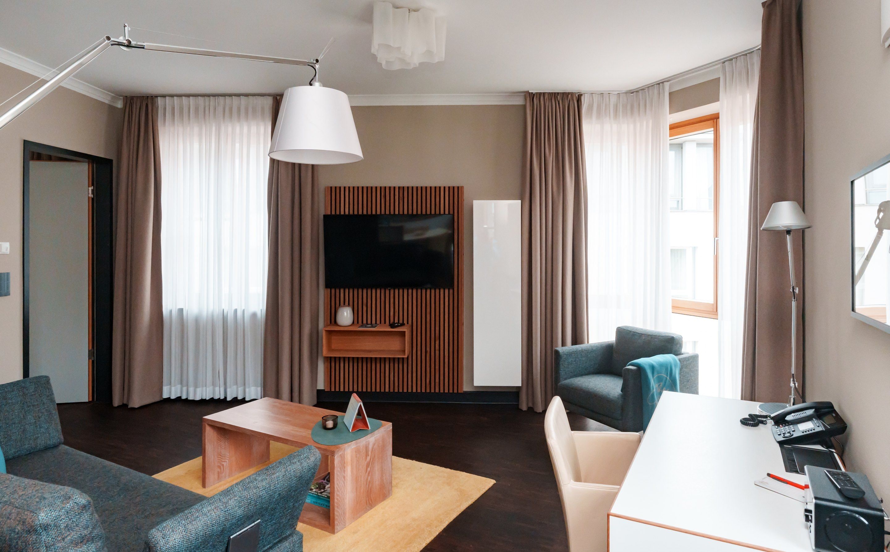 A modern hotel room with a flat-screen TV, a wooden accent wall, a sofa, a coffee table, a desk with a phone, and a lamp.