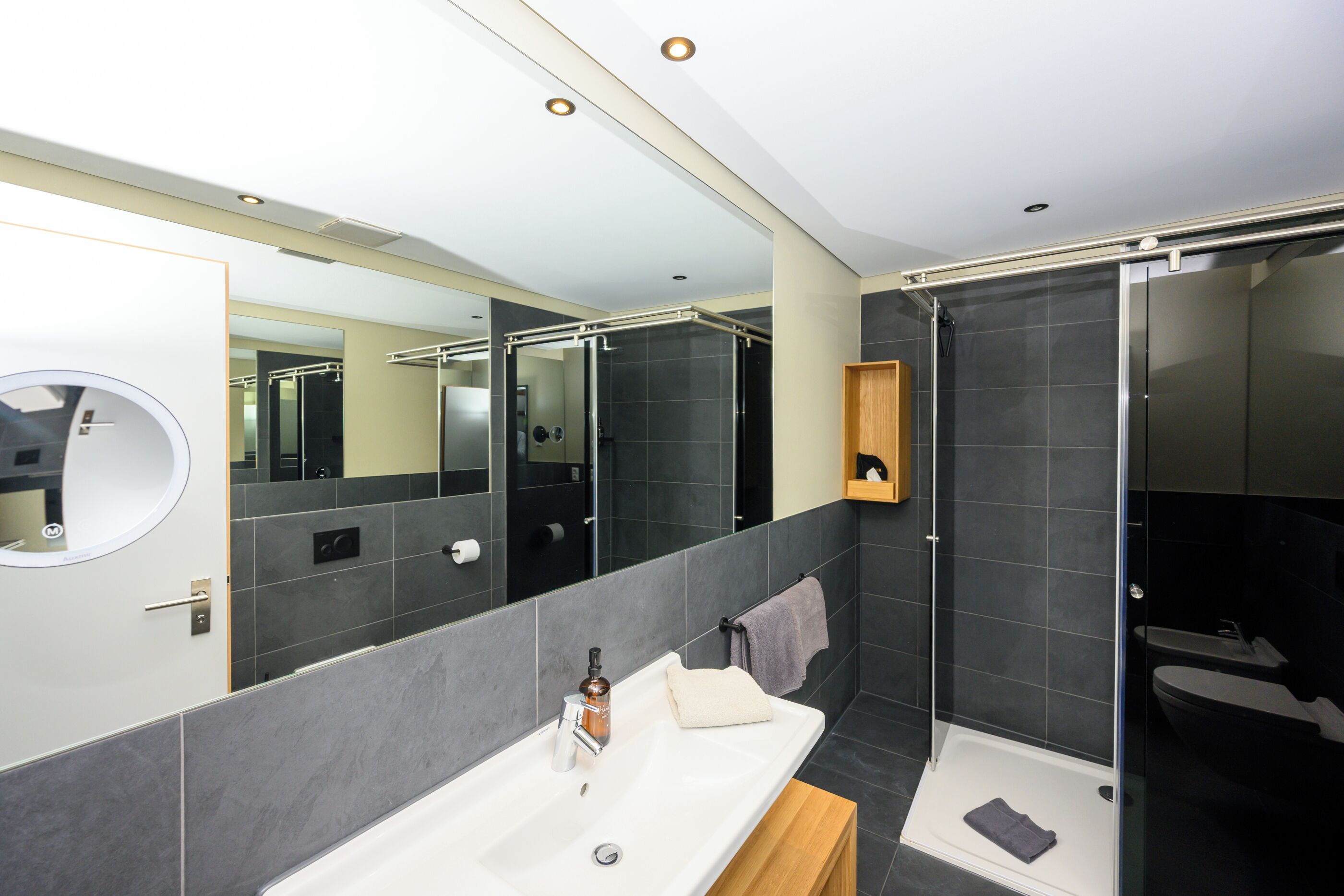 Un baño moderno con un espejo grande, un lavamanos blanco, una zona de ducha y un estante de madera.
