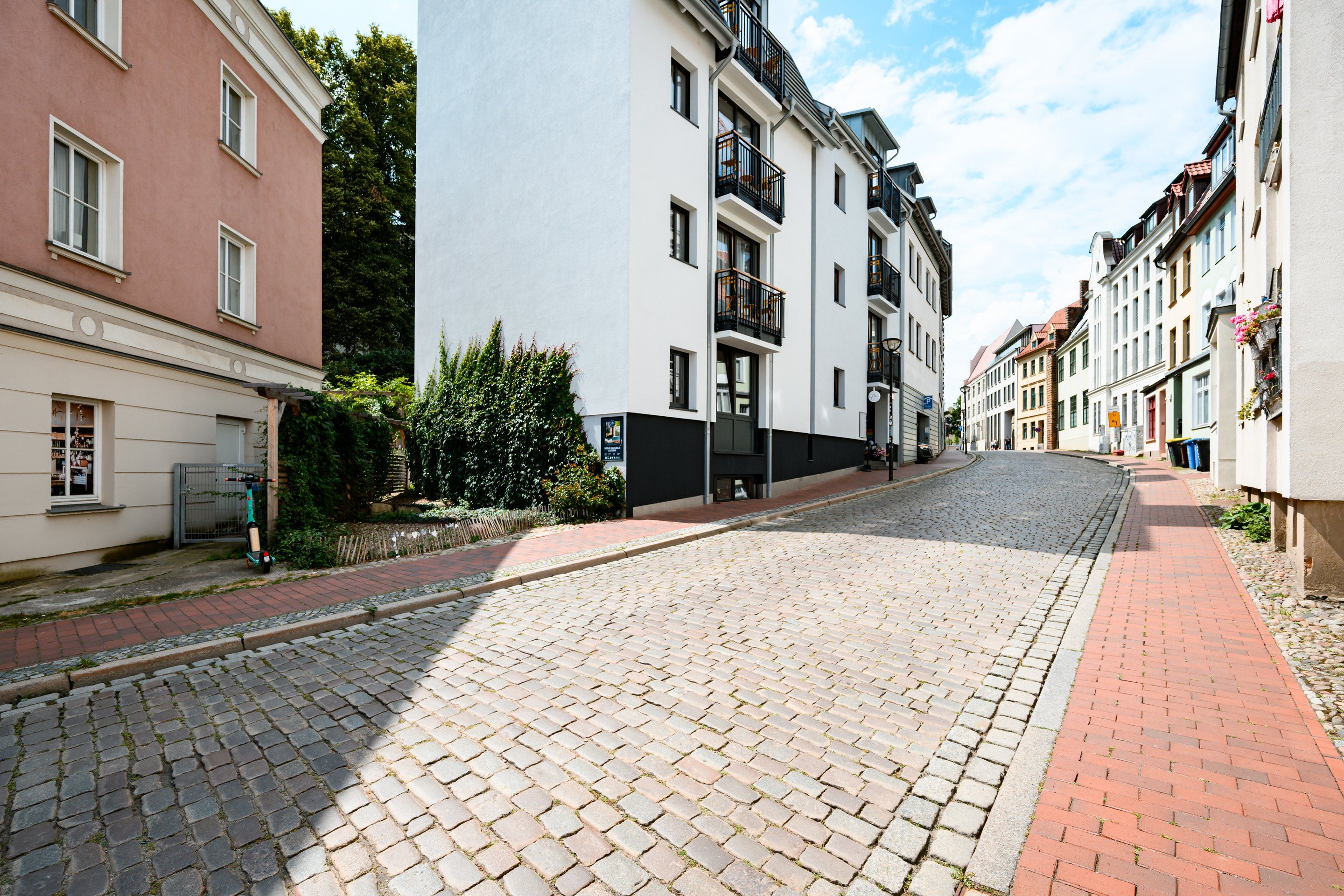 A cobblestone street lined with multi-story buildings, some with balconies and greenery.