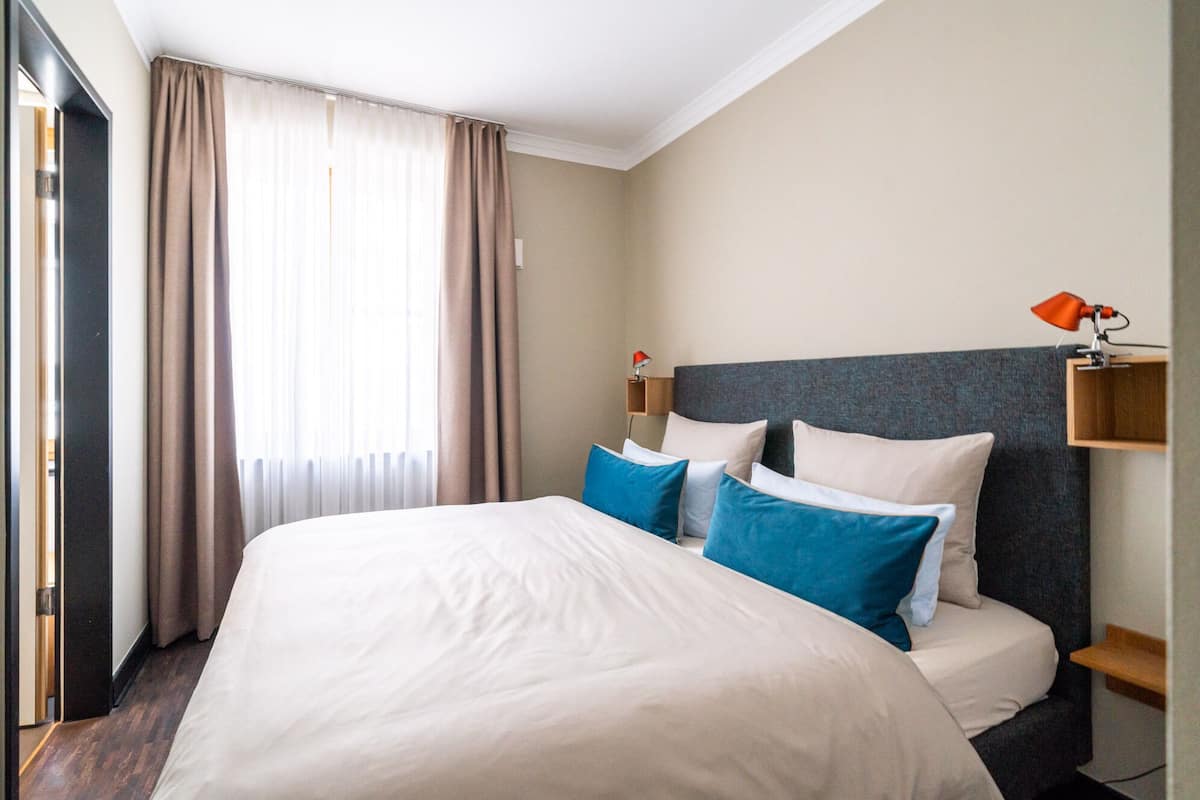 A hotel room with a large bed, two bedside tables, a nightstand, a lamp, and a window with curtains.