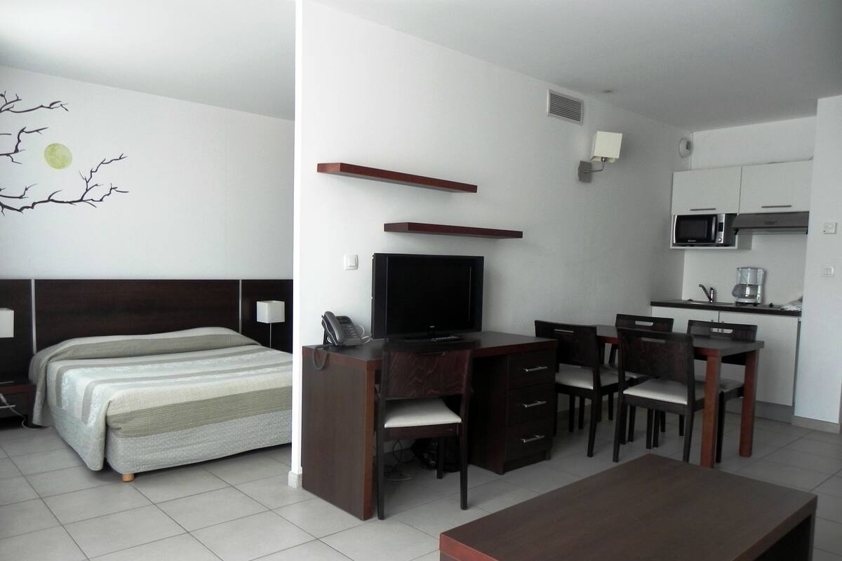 A hotel room with a bed, a desk with a television, a chair, a small table, and a kitchenette.