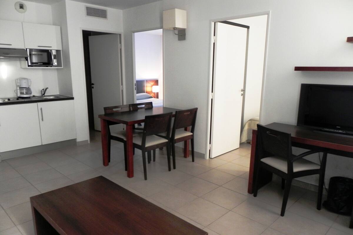 A compact living space with a kitchenette, a dining area with a table and chairs, a TV, and a bench.