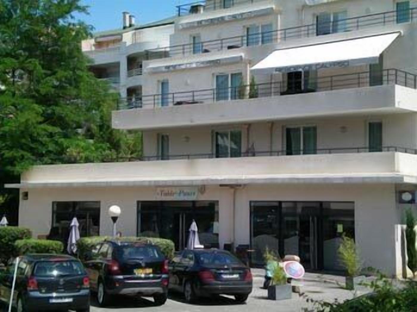 Un hôtel à plusieurs étages au design moderne, doté de balcons à chaque niveau, d'une entrée principale avec une enseigne et d'un parking où sont stationnées plusieurs voitures.