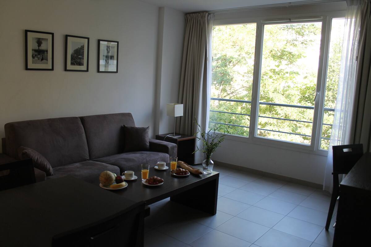 A modern living room with a sofa, coffee table, and dining area. There are framed pictures on the wall, a window with curtains, and a plant on the table.