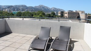 A rooftop terrace with two wicker lounge chairs, a tiled floor, and a view of buildings and mountains in the background. - Residence Calypso (Marseille)