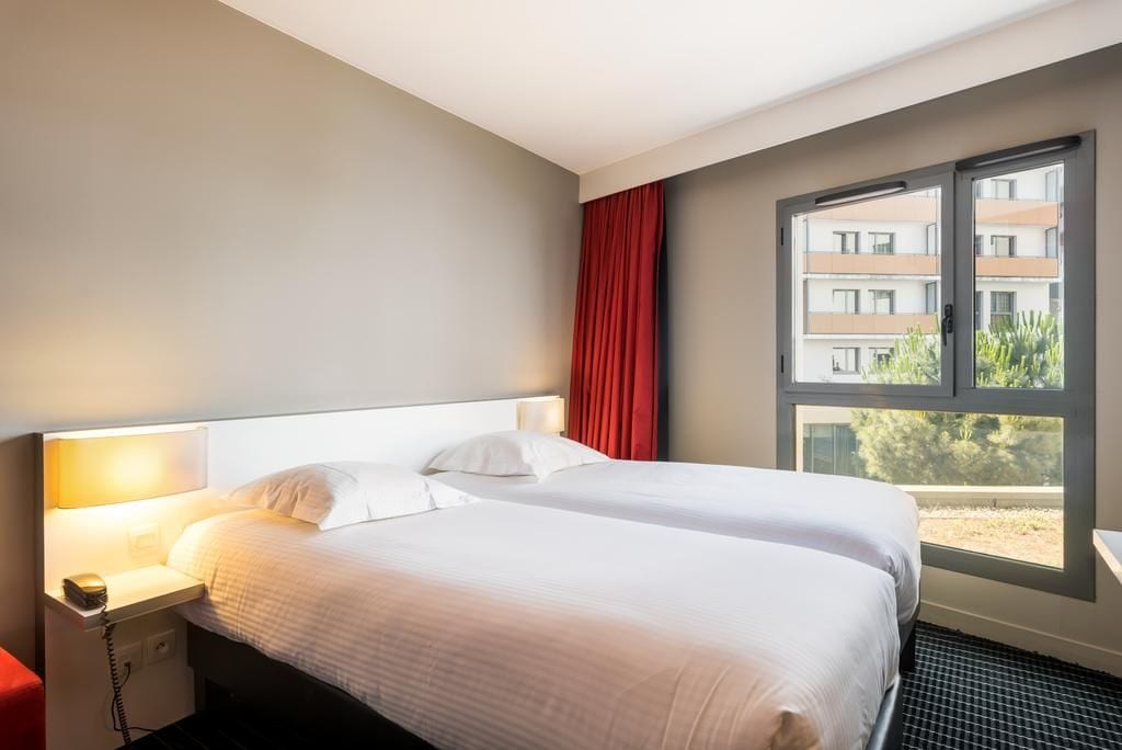 A hotel room with a bed, a nightstand, a lamp, a window with a view of buildings, and a red curtain.