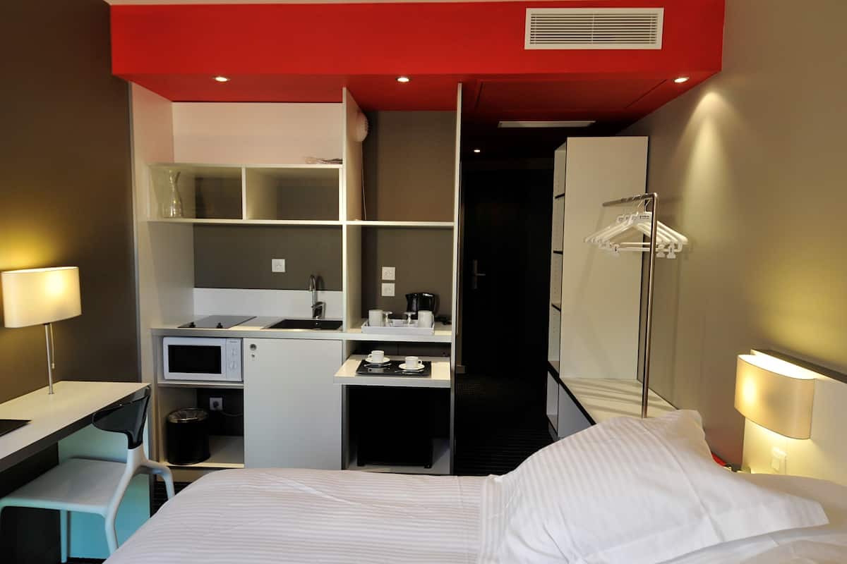 Habitación de hotel pequeña con cama, escritorio, microondas, cafetera y armario.