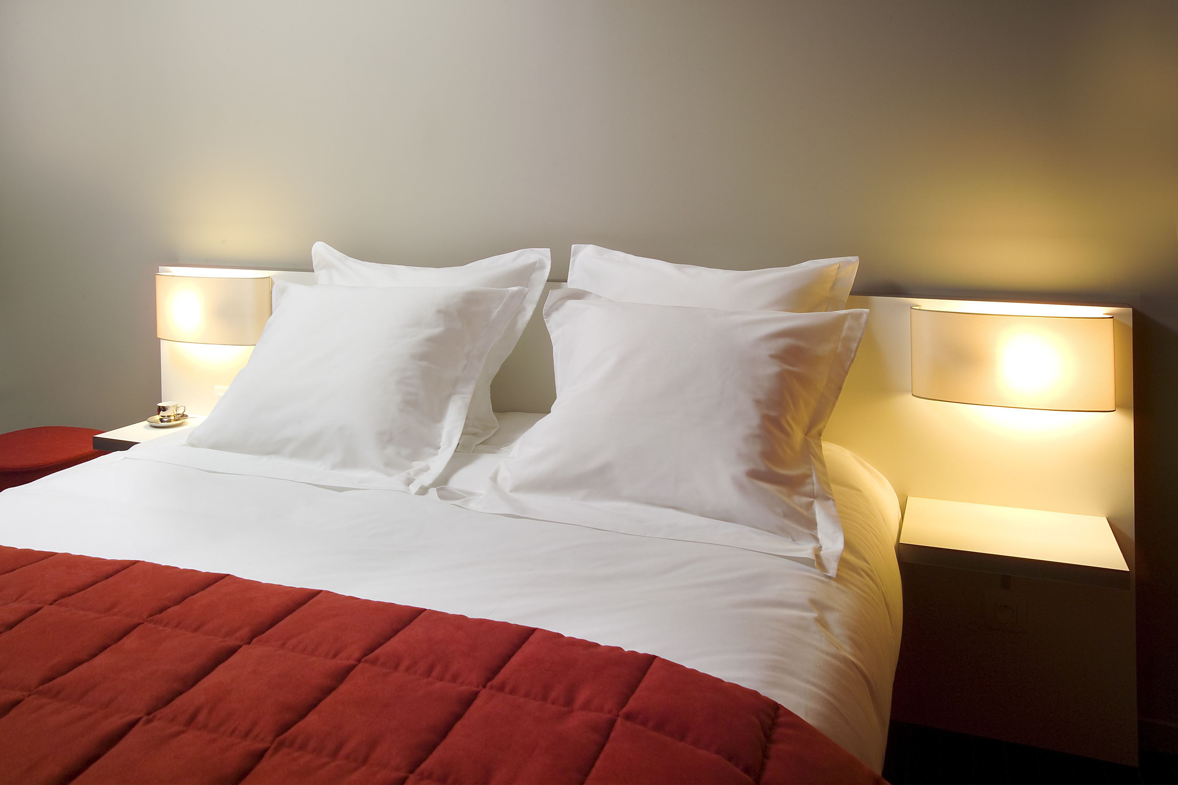 Ein ordentlich bezogenes Bett mit weißen Laken und einer roten gesteppten Tagesdecke. Zwei Nachttischlampen mit weißen Lampenschirmen und ein kleiner Tisch mit Tasse und Untertasse neben dem Bett.