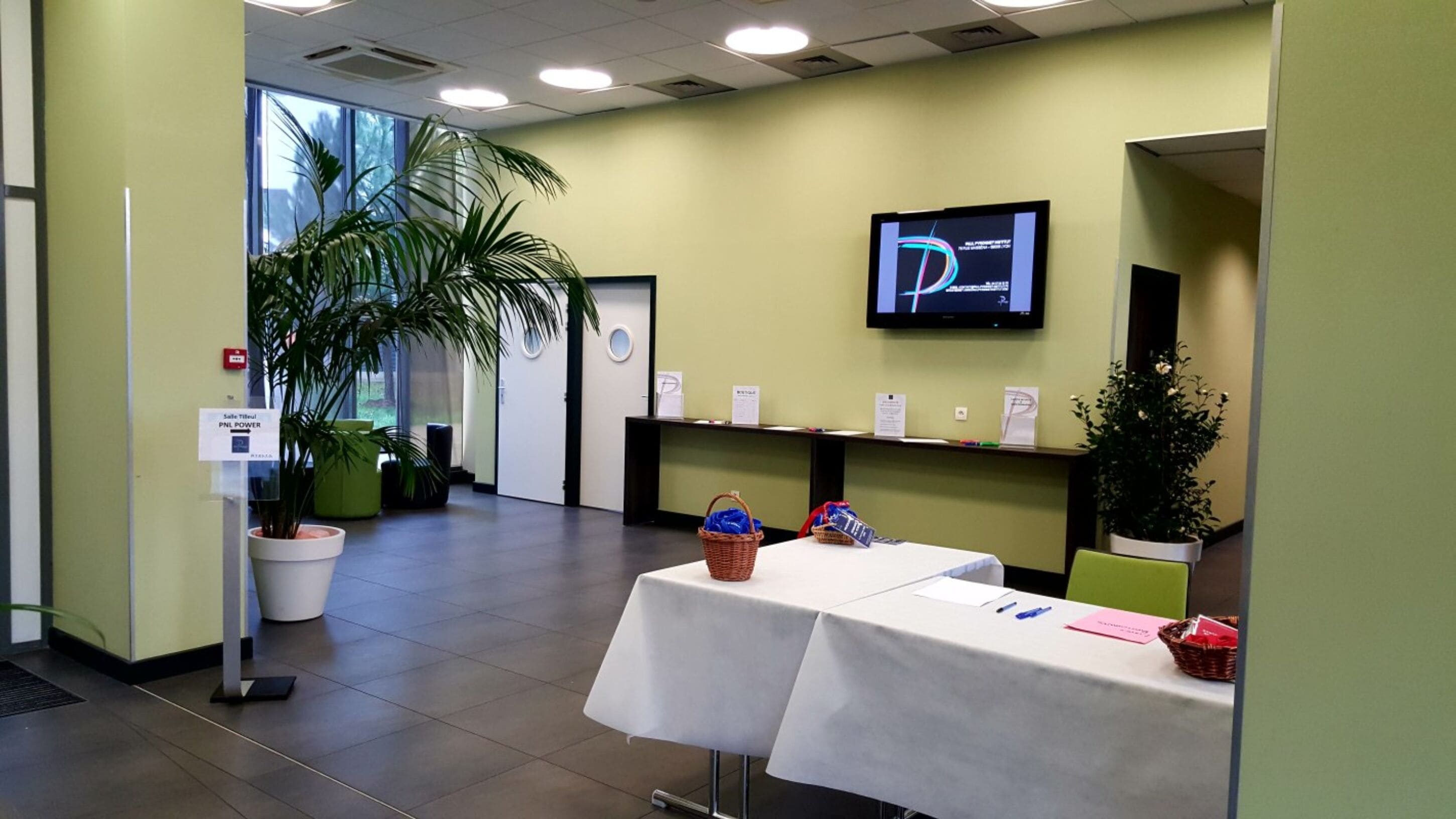 A reception area with a reception desk, a television screen, potted plants, and a sign indicating the location.