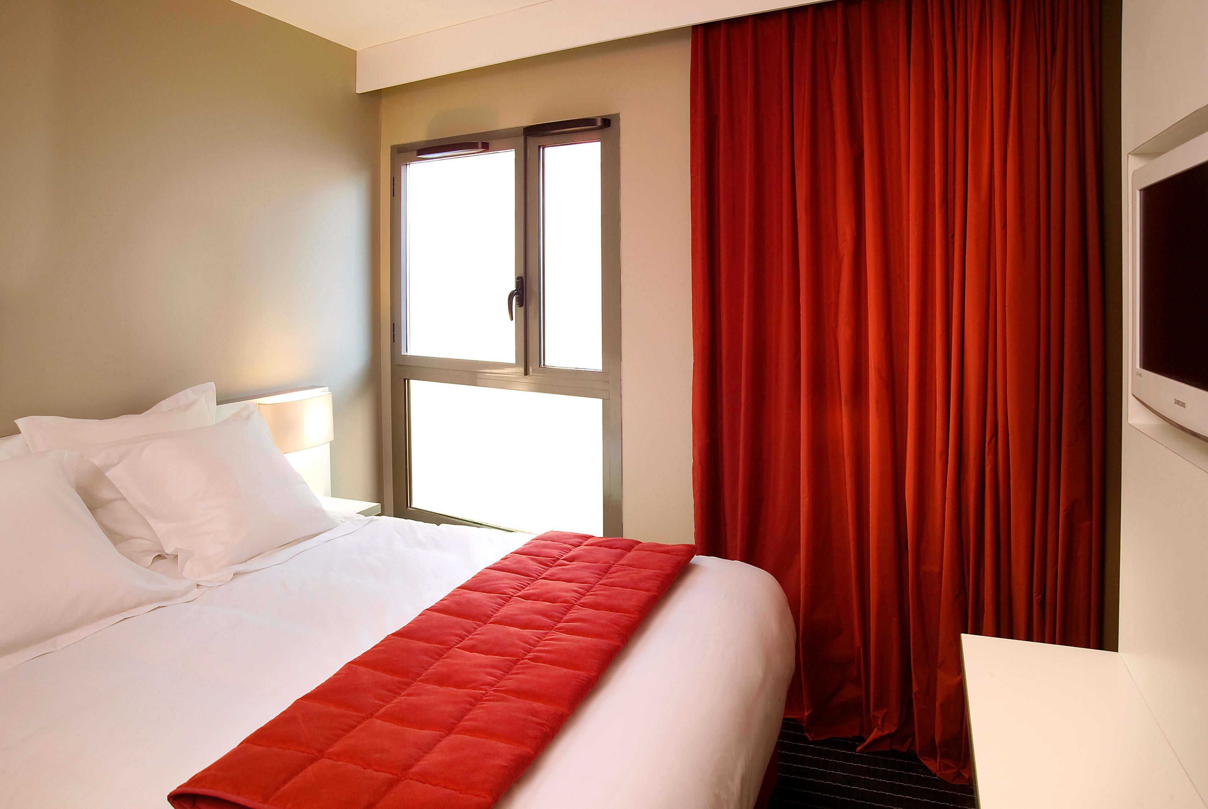 A hotel room with a bed, white bedding, a red blanket, a window with curtains, and a wall-mounted TV.