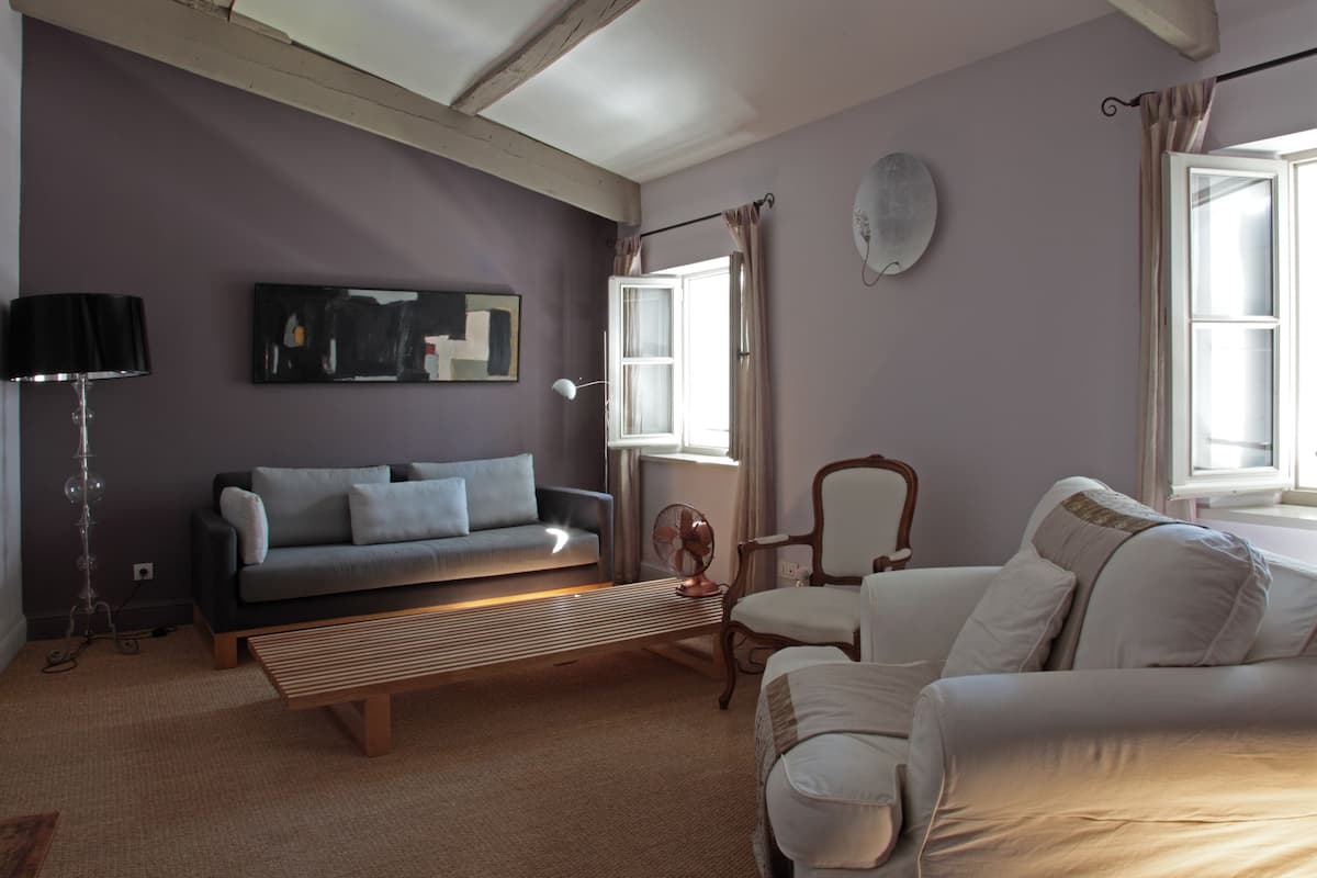 Ein Wohnzimmer mit Sofa, Sessel und einem Ventilator. Es gibt ein Fenster mit Vorhängen und eine Wandleuchte.