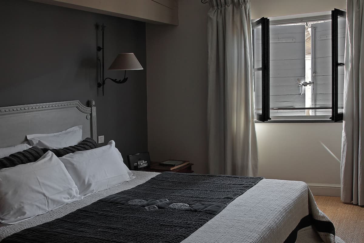 Ein ordentlich bezogenes Bett mit weißen und dunklen Kissen, eine Wandlampe und ein Fenster mit Vorhängen.