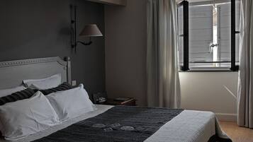 A neatly made bed with white and dark pillows, a lamp on the wall, and a window with curtains.