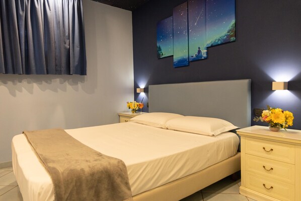 A neatly made bed with a beige blanket, a bedside table with a vase of flowers, and a wall-mounted night light. - Pompei Valley Boutique Hotel (Pompei)