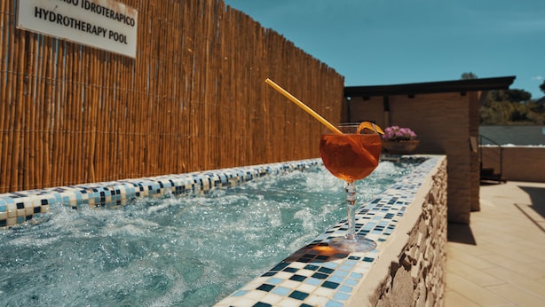 Uma piscina de hidroterapia com uma placa indicando "PERCORSO IDROTHERAPICO HYDROTHERAPY POOL" e um copo de bebida com canudo na borda da piscina.