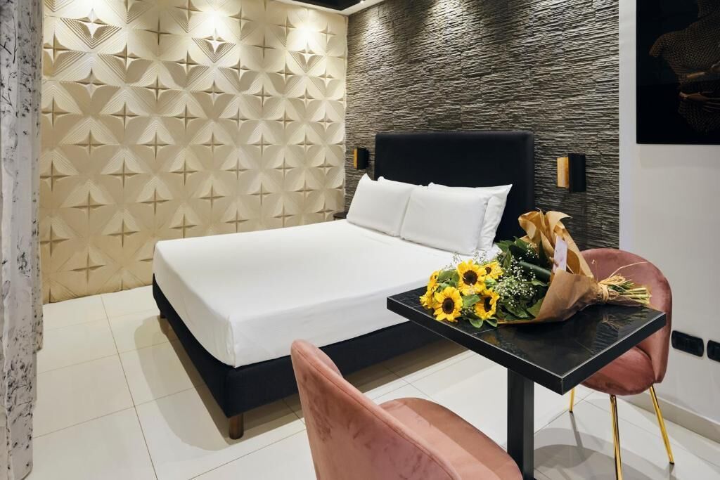 Une chambre d’hôtel moderne avec un lit, une table ornée d’un bouquet de tournesols et une chaise rose.