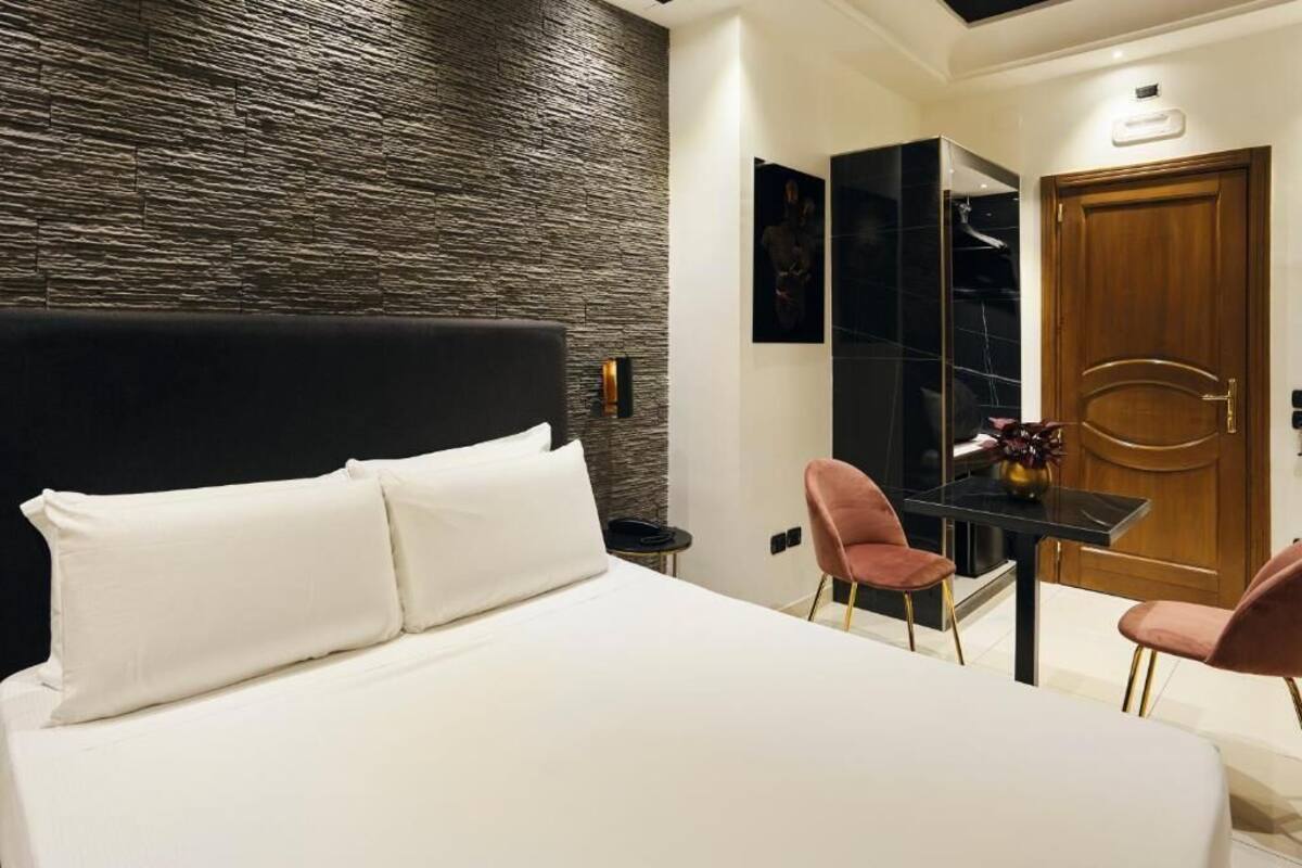 Una camera d'albergo moderna con un letto grande, un tavolino con due sedie, una porta in legno e un parete con texture.